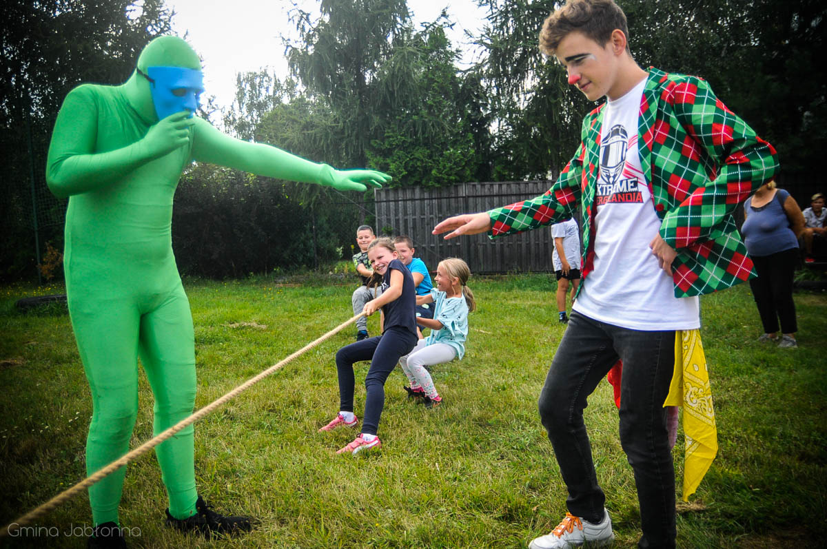 Człowiek w zielonym, obcisłym kombinezonie oraz klaun nadzorują przebieg konkursu w przeciąganiu liny. 